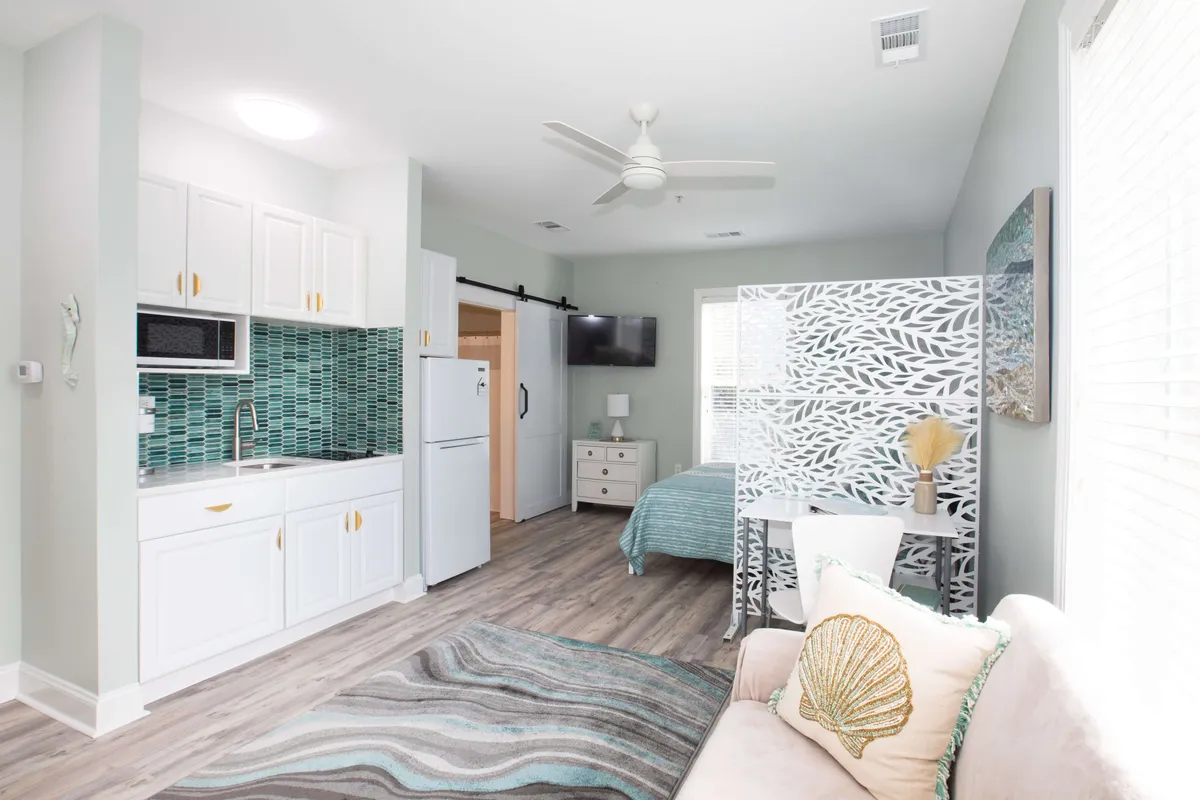 A view of the studio’s open-plan layout including a kitchenette, separating screen, couch, and bed. Warm, natural light enters through the windows, and the space feels inviting and functional, supporting activities such as relaxing, reading, and enjoying a quiet meal.