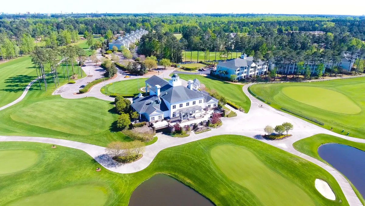 A sweeping exterior view of the golf clubhouse surrounded by lush green lawns and an expansive golf course, with tranquil ponds dotting the landscape. Outdoor dining areas and pathways invite relaxation and community gatherings, all set under warm, inviting light.