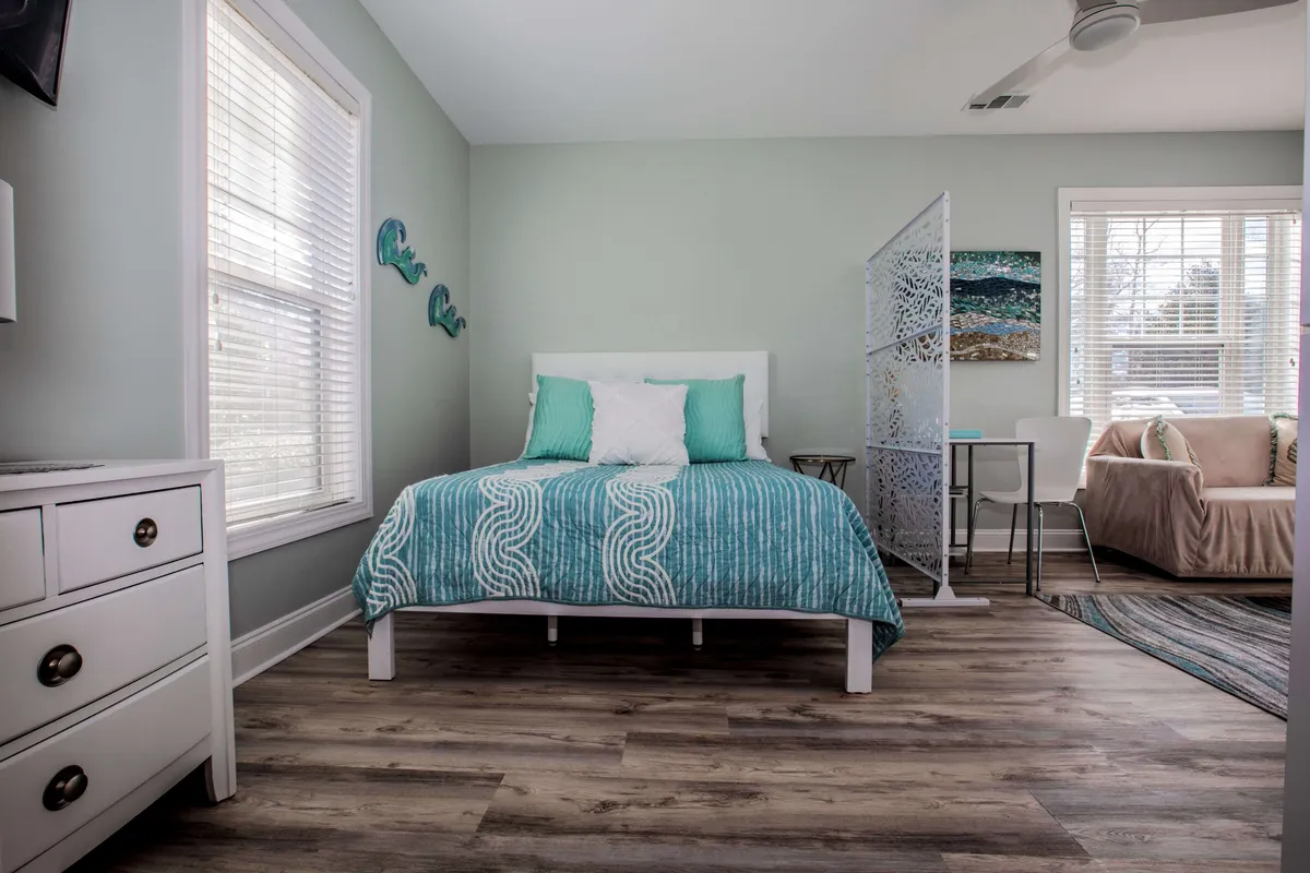 A tranquil studio corner featuring a bed with teal and white bedding, decorative wall art of waves, a partition screen, and a comfortable seating area. The space is bathed in soft natural light, creating an inviting atmosphere that encourages relaxation and quiet mornings.