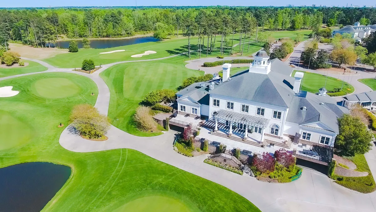 An elegant exterior shot capturing the clubhouse set against manicured fairways, mature trees, and sparkling water features. The scene conveys tranquility and luxury, with glimpses of guests enjoying the picturesque setting by the water.