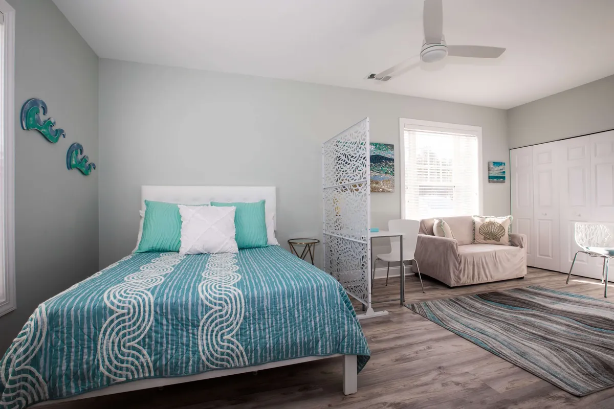 The image showcases a stylish, sunlit studio interior featuring a plush bed with patterned bedding, a decorative partition that subtly divides the sleeping area from a comfortable seating nook with a sofa and chair. The setting evokes relaxation, with glimpses of a table perfect for morning coffee or leisurely reading, creating a cozy, serene ambiance.