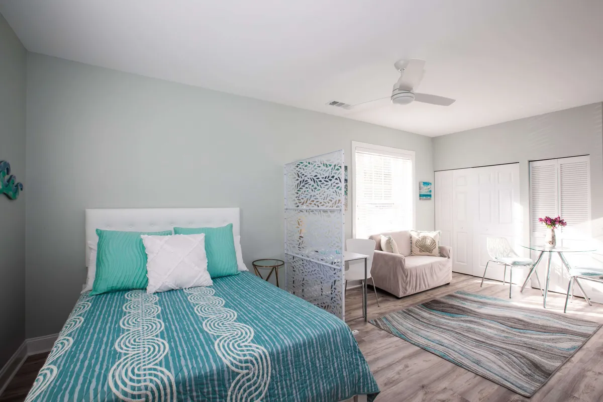 A serene studio bedroom with turquoise bedding, a decorative screen, and a cozy seating area with a glass table. Morning sunlight filters through sheer curtains, creating a welcoming and restful vibe—perfect for relaxation between shifts.