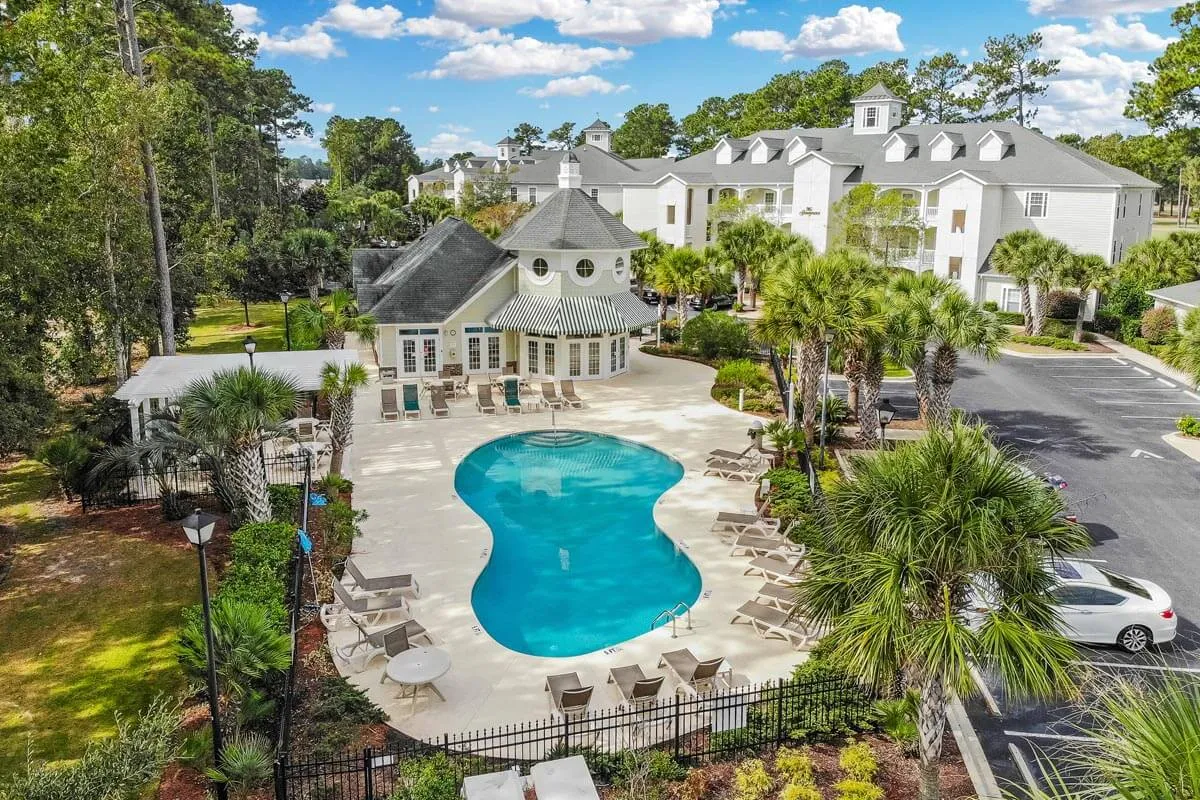 This image features a sparkling outdoor swimming pool surrounded by palm trees and a clubhouse, evoking a sense of leisure and tranquility. The setting is perfect for enjoying a refreshing swim or relaxing poolside with your partner, with sunset light reflecting off the water to create a romantic ambiance.