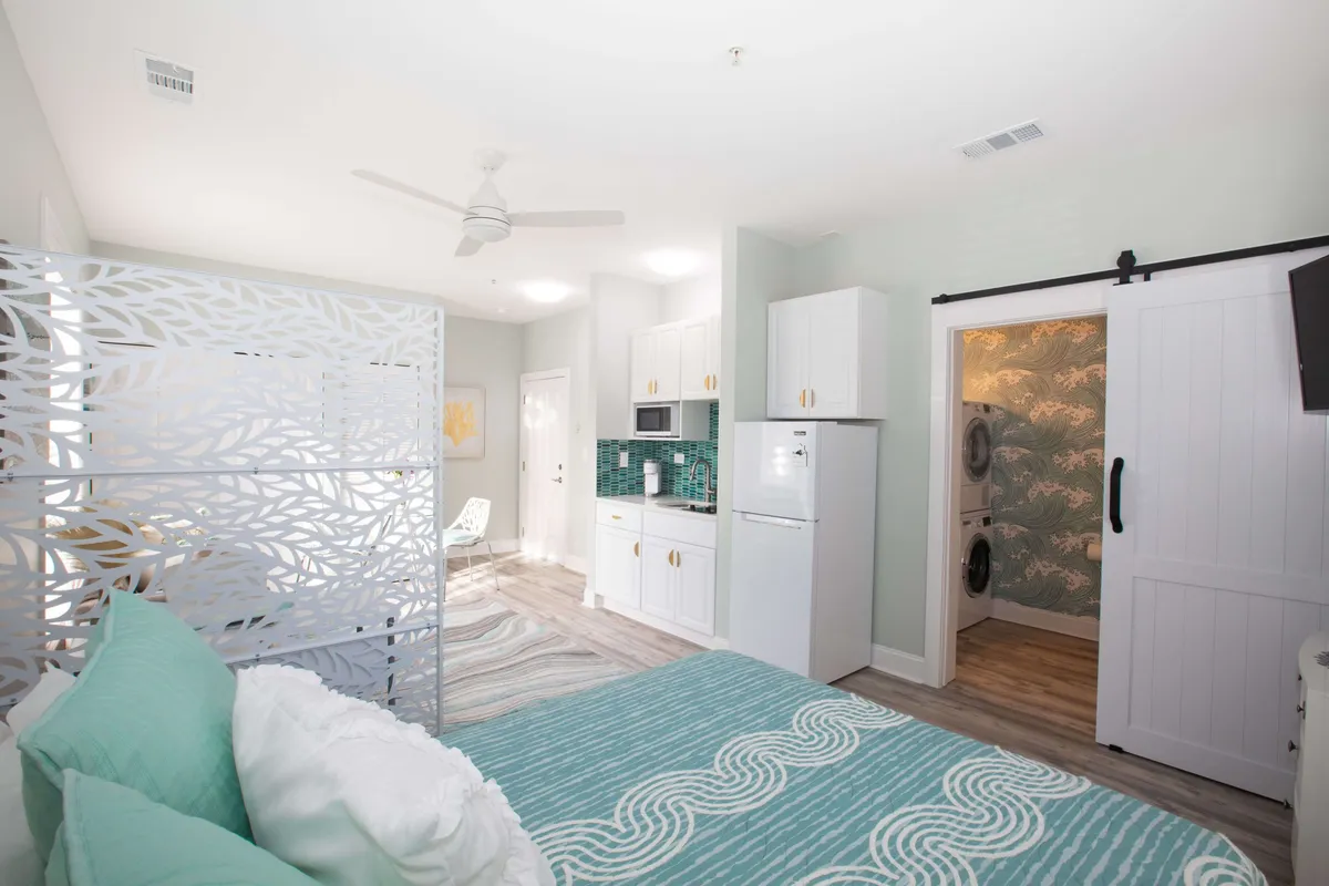 This image shows a modern studio layout with a bed featuring turquoise bedding, a stylish room divider, kitchenette, and a washer/dryer unit. Sunlight streams through the window, brightening both the sleeping and living areas. The kitchenette and laundry units suggest comfort and utility for longer stays, supporting the 'work and wellness' narrative.