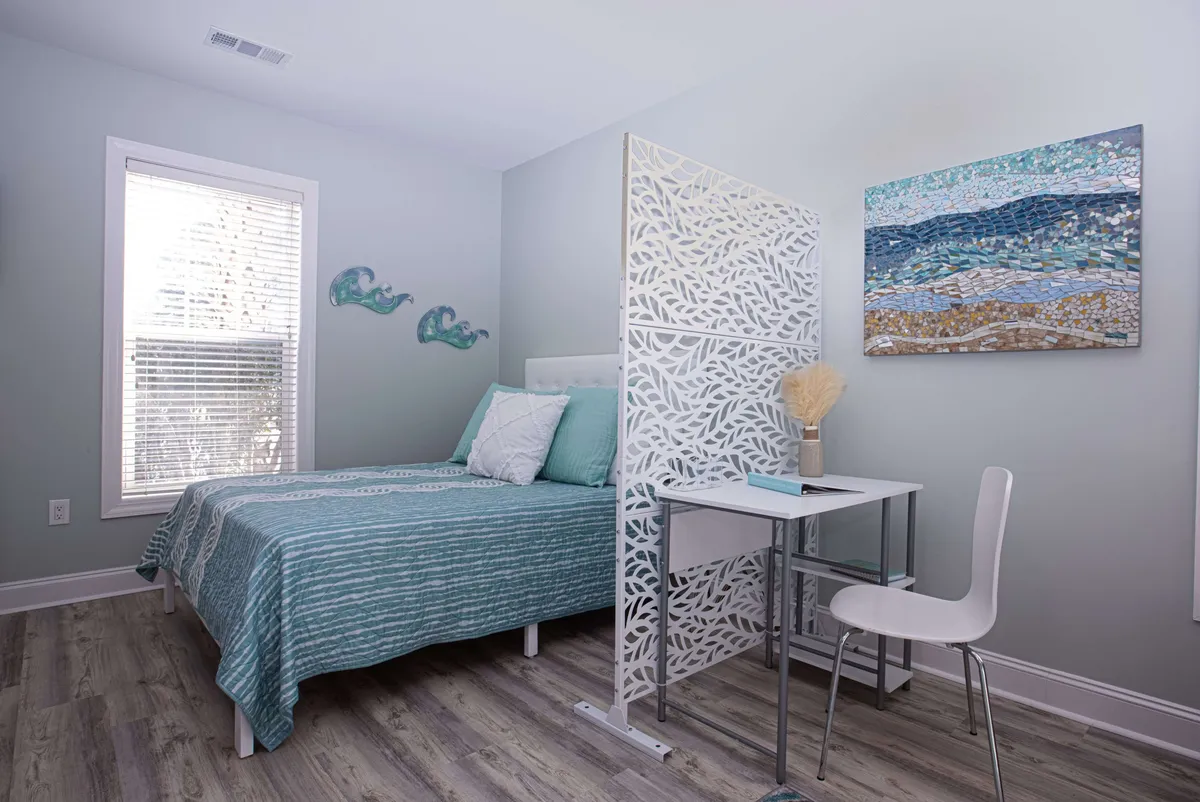 This image depicts a serene studio space featuring a bed with teal bedding, a decorative room divider, and a neatly arranged desk and chair positioned near a window. The calming decor and abundance of natural light create an inviting setting for both rest and focused work, while the visible desk highlights the workspace aspect.