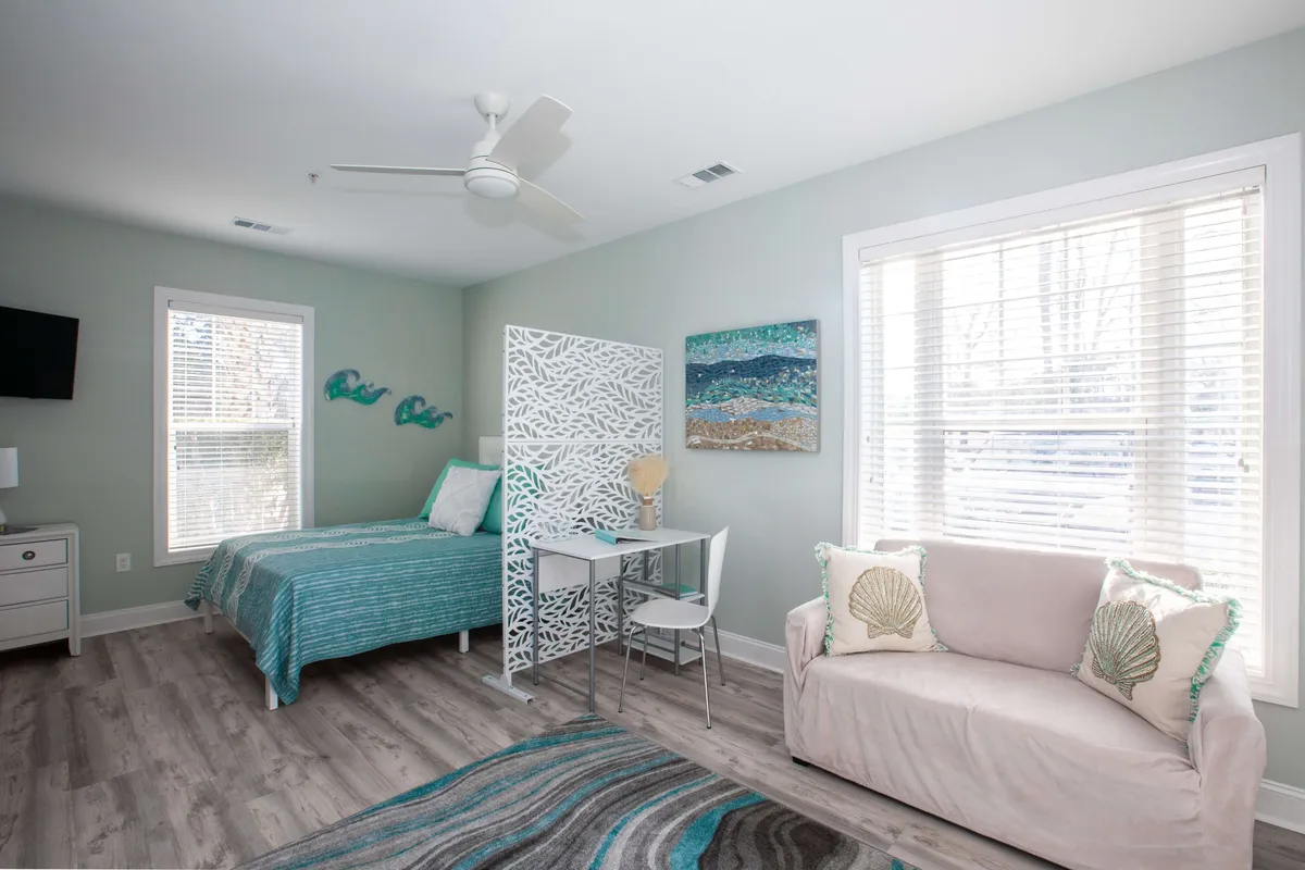 A bright studio space featuring a bed with teal and white bedding, a decorative room divider, artwork depicting an ocean scene, and a comfortable sofa adorned with seashell-themed cushions. Natural light streams in through the window, enhancing the tranquil and coastal atmosphere. There is also a small desk area, perfect for reading or working.